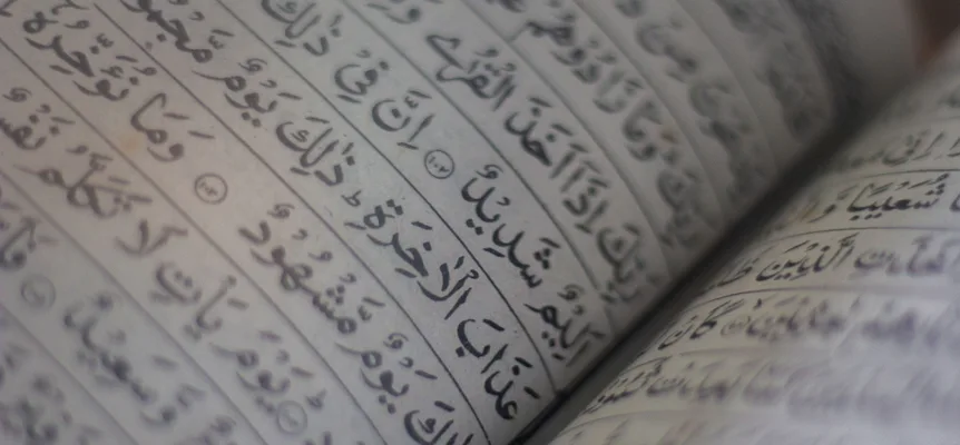 Close-up of the curved pages of an open book printed in fully vowelled Arabic script, with round verse markers between the words