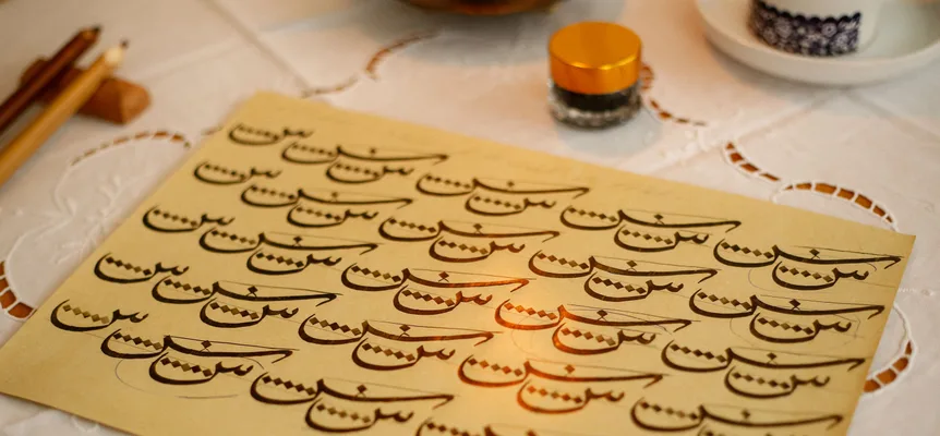 A cream practice sheet on a white lace tablecloth, filled with rows of one Arabic letter stroke in black ink with small diamond dots beside each, two reed pens and an ink pot nearby