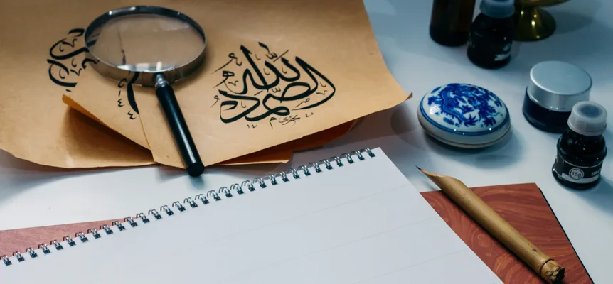 Sheets of Arabic calligraphy on a desk beside ink bottles, a magnifying glass and a blank notepad