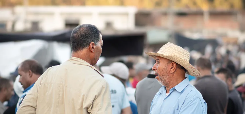 Two men talking at a crowded outdoor market, one in a beige jacket with his back half turned, the other in a straw hat and a light blue shirt