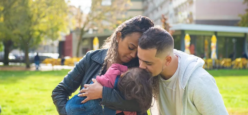 A woman holds a small child in a city park while a man leans in to kiss the child's head