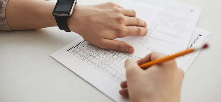 A hand holding an orange pencil fills in a multiple-choice bubble sheet while the other hand, wearing a black smartwatch, rests on the test booklet