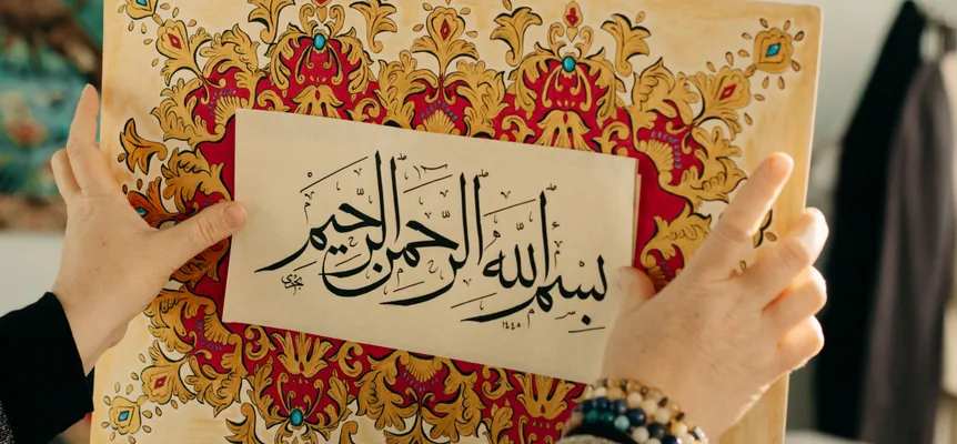 Two hands hold up a sheet of black Arabic calligraphy in front of a board painted with a gold, red and turquoise floral border