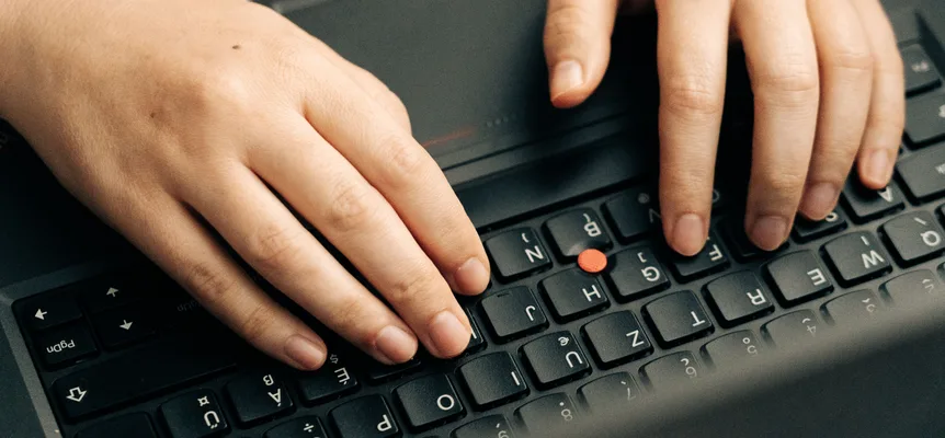 Two hands rest on the keys of a black laptop with a red pointing stick, photographed from behind the screen so the key letters appear upside down