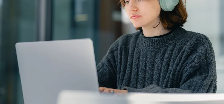 A young woman in a grey knit sweater and mint-green headphones looking at a silver laptop on a table
