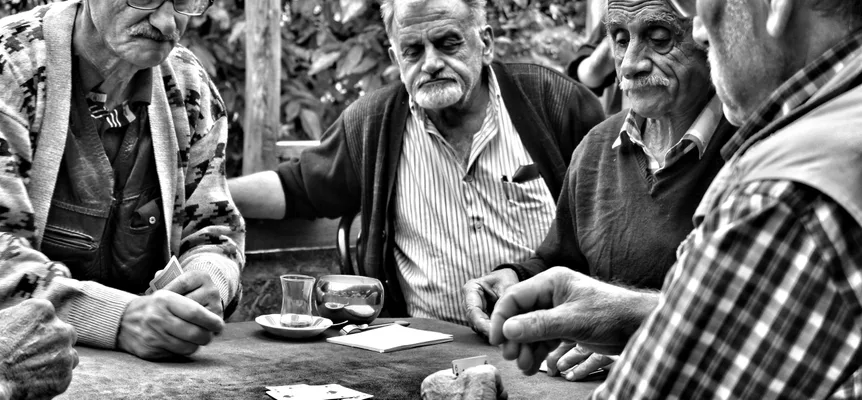 Black-and-white photo of four older men playing cards at an outdoor café table with a glass of tea
