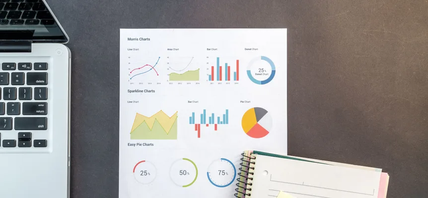 A printed sheet of sample line, bar, pie and donut charts on a dark desk, between an open laptop and a spiral notebook