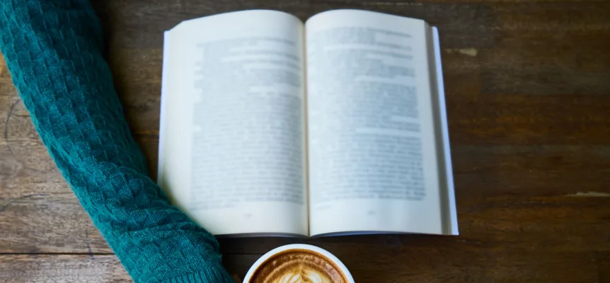 An open paperback on a wooden table next to a latte with leaf-pattern foam
