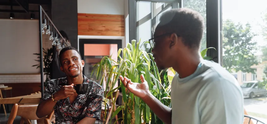 Two young men chatting in a cafe by a large window, one holding a black coffee cup while the other gestures