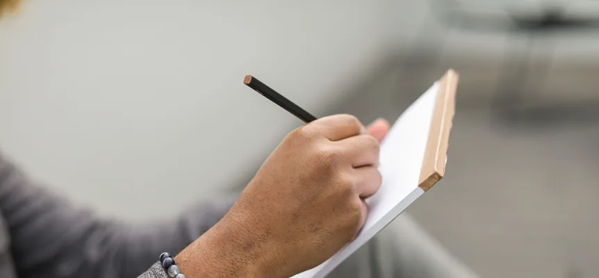 A hand wearing a beaded bracelet writes with a black pencil on a white notepad