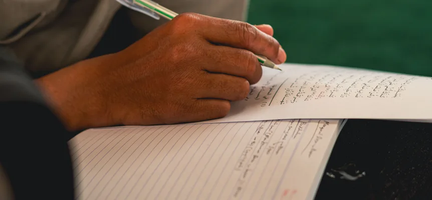 A hand holding a green pen writes lines of Arabic script on a sheet of paper laid over a lined notebook