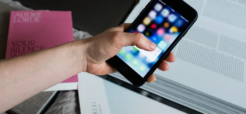 A hand holds a smartphone over an open book, thumb pressing an app icon on the home screen