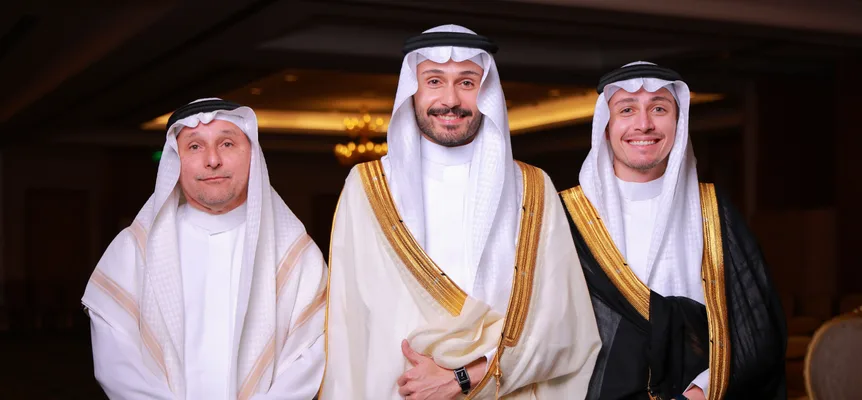 Three men in white thobes, white ghutras and black ʿiqals; two wear gold-trimmed bishts, one cream and one black, in a hall with a chandelier