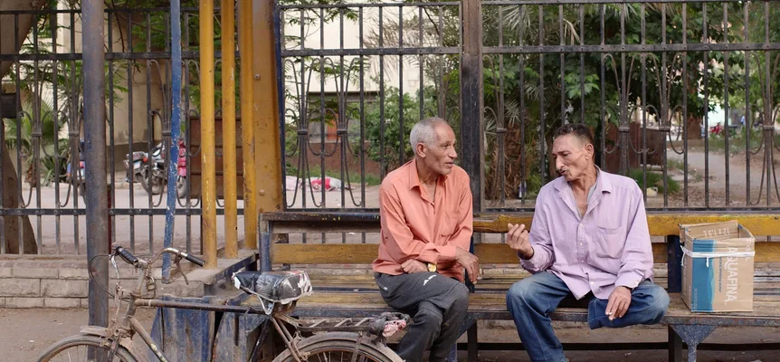 Two older men talking on a wooden bench beside an iron fence on a Cairo street, with a bicycle parked in front