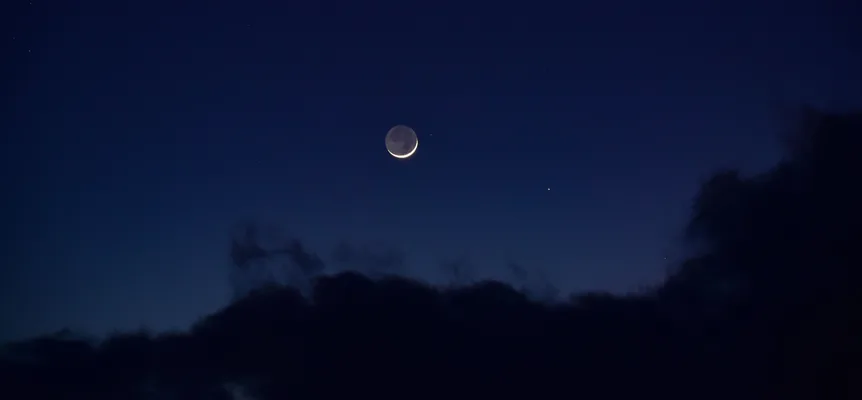 A thin crescent moon in a dark blue evening sky above a bank of dark clouds