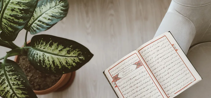 An open copy of the Quran with red-bordered pages resting on the arm of a grey armchair, beside a potted plant on a pale wooden floor