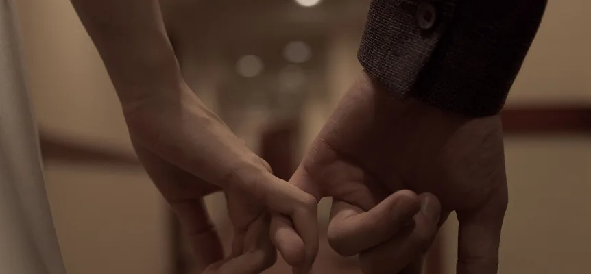 Two hands, one larger than the other, linked by their little fingers against a blurred, warmly lit room