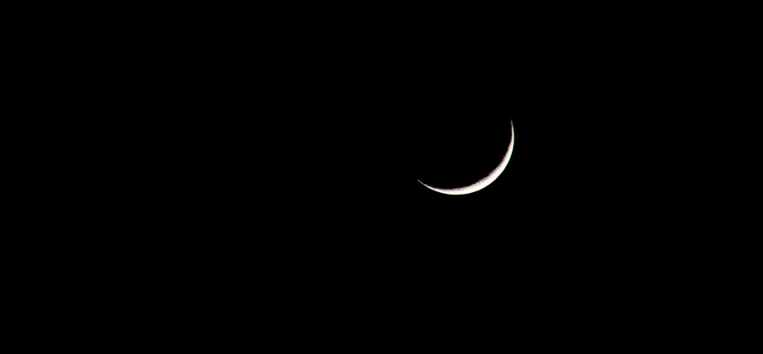A thin crescent moon, lit along its lower edge, alone in a black night sky