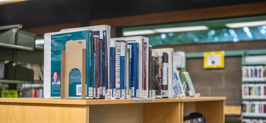 A public library shelf filled with hardback books and DVD cases under warm light