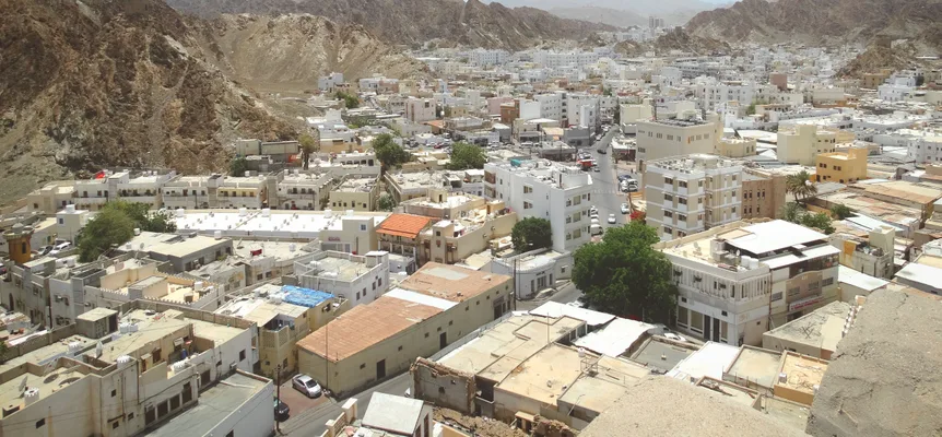 Low white and sand-coloured buildings of a Muscat neighbourhood packed into a valley between bare brown hills