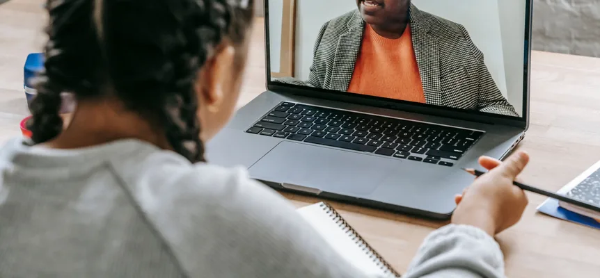 A student with braided hair, seen from behind, holds a pencil while a teacher in an orange top and checked jacket speaks on a laptop video call