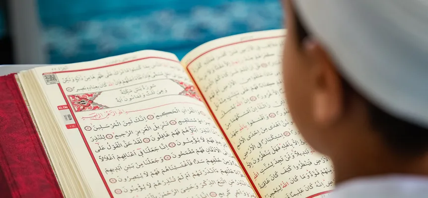 Über die Schulter eines Jungen mit weißer Kappe gesehen: ein aufgeschlagener Koran mit rot umrandeten Seiten und arabischem Text