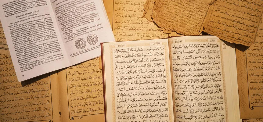 Arap harfleriyle yazılmış, sararmış, dağınık el yazması sayfaların üzerinde duran açık bir basılı Kur'an ve yanında Kiril harfleriyle basılmış açık bir kitap