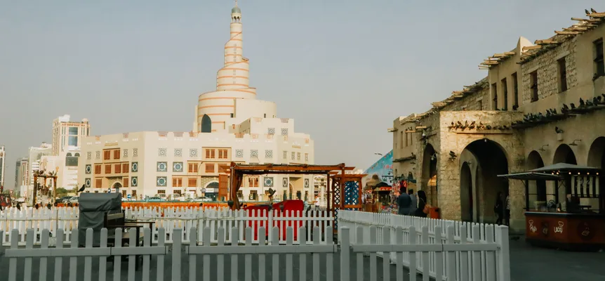 A white spiral minaret above a cream building, with Souq Waqif's stone arcades and a white picket fence in front