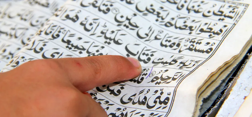 A finger rests under a line of black Arabic script on a worn Quran page printed without color coding