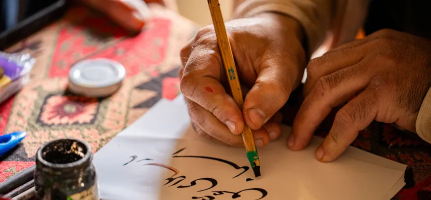 Two hands writing Arabic-script calligraphy in black ink with a wooden reed pen on white paper, an open ink pot beside it on a red patterned rug